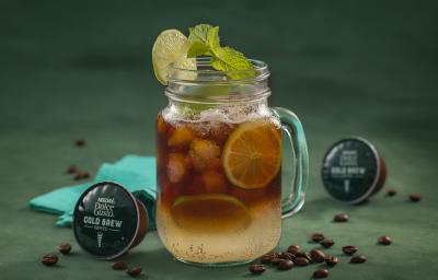 Fotografia em tons de verde de uma bancada verde, sobre ela um copo de vidro com cold brew e limões. Ao lado capsulas de Dolce Gusto Cold Brew e um paninho azul.