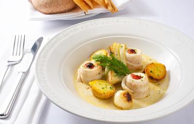 Fotografia em tom claro de linguado enrolado e batatas com um creme por baixo dentro de um prato branco fundo. Ao lado, talheres apoiados sobre uma toalha branca em uma mesa também na cor branca.