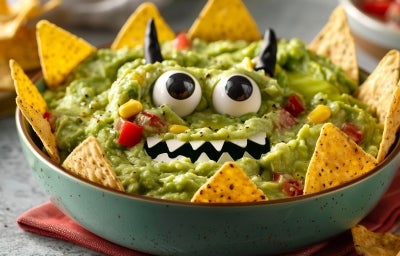 Fotografia mostra guacamole cremoso em formato de monstro verde, com olhos de ovo e azeitona, dentes de queijo e nachos crocantes formando espinhos ao redor.