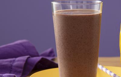 Fotografia de um copo de vidro grande com um smoothie de açaí apoiado em um prato amarelo raso com um canudo branco e roxo. Ao lado tem uma embalagem de ninho proteen, sobre uma mesa de madeira. Fotografia de um copo de vidro grande com um smoothie de açaí apoiado em um prato amarelo raso com um canudo branco e roxo. Ao lado tem uma embalagem de ninho proteen, sobre uma mesa de madeira.