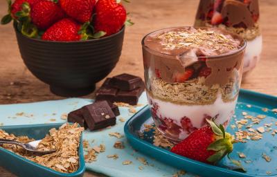 Fotografia em tons de vermelho em uma bancada de madeira, uma tábua azul, um recipiente quadrado azul com um copo de vidro com o creme branco, com Neston 3 Cereais, creme de chocolate e morangos. Fotografia em tons de vermelho em uma bancada de madeira, uma tábua azul, um recipiente quadrado azul com um copo de vidro com o creme branco, com Neston 3 Cereais, creme de chocolate e morangos.