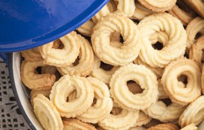 Fotografia em tom claro, com biscoitos amanteigados em formato redondo, modelado com bico de confeitar trançado, dentro de um refratário. No canto superior esquerdo da foto, uma tampa azul. Fotografia em tom claro, com biscoitos amanteigados em formato redondo, modelado com bico de confeitar trançado, dentro de um refratário. No canto superior esquerdo da foto, uma tampa azul.