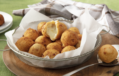 Foto da receita de bolinho de batata servida em diversos bolinho dentro de uma peneira de metal coberta com guardanapos sobre uma tábua de madeira com uma colher prateada ao lado com um bolinho. Ao fundo um paninho cinza quadriculado