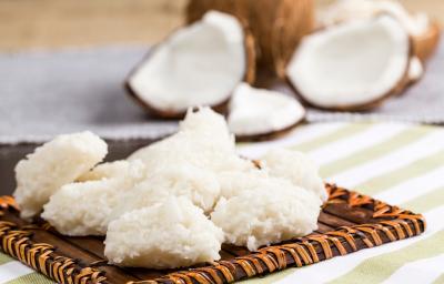 Foto da receita de Cocada Zero Lactose. Observa-se as cocadas dispostas em uma tábua e atrás é possível ver os cocos abertos. Foto da receita de Cocada Zero Lactose. Observa-se as cocadas dispostas em uma tábua e atrás é possível ver os cocos abertos.