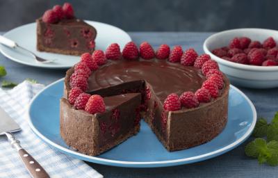 Fotografia em tons de vermelho em uma bancada de madeira azul, um pano azul quadriculado com uma espátula para bolo em cima dele. Ao centro, um prato azul redondo raso com a torta cremosa de chocolate com framboesas. Ao fundo, um prato com uma fatia. Fotografia em tons de vermelho em uma bancada de madeira azul, um pano azul quadriculado com uma espátula para bolo em cima dele. Ao centro, um prato azul redondo raso com a torta cremosa de chocolate com framboesas. Ao fundo, um prato com uma fatia.