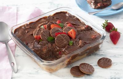 Fotografia em tons de vermelho em uma bancada de madeira clara, um pano branco com listras vermelhas, um refratário quadrado de vidro com a lasanha doce de chocolate com morangos e biscoito calipso dentro dele. Ao fundo, um prato azul com fatia do doce. Fotografia em tons de vermelho em uma bancada de madeira clara, um pano branco com listras vermelhas, um refratário quadrado de vidro com a lasanha doce de chocolate com morangos e biscoito calipso dentro dele. Ao fundo, um prato azul com fatia do doce.