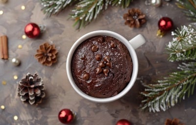 Caneca branca cheia de bolo de chocolate, decorada com gotas de chocolate e açúcar de confeiteiro. A caneca está cercada por enfeites natalinos, como ramos de pinheiro, pinhas e ornamentos vermelhos e prateados.