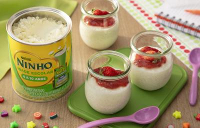 Fotografia de três recipientes de vidro pequenos com pudim de tapioca de ninho um creme de morango por cima. Dois desses recipientes estão sobre um apoio verde retangular, ao lado de uma colher roxa pequena e de uma lata de ninho escolar.