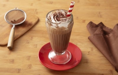 Fotografia em tons claros de um copo grande de vidro, de milkshake, com a bebida de nescau, finalizada com chantilly e nescau. O copo está sobre um mini prato vermelho, apoiado em uma mesa de madeira clara. Fotografia em tons claros de um copo grande de vidro, de milkshake, com a bebida de nescau, finalizada com chantilly e nescau. O copo está sobre um mini prato vermelho, apoiado em uma mesa de madeira clara.