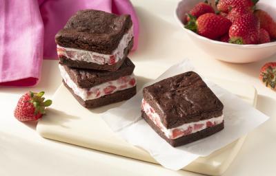 Imagem aproximada da receita de Sanduíche de Brownie com Sorvete, em quadrados marrons, recheados com um creme claro com pedaços de morango, todos sobre uma tábua clara em uma bancada decorada com mais morangos e um tecido em tom rosa Imagem aproximada da receita de Sanduíche de Brownie com Sorvete, em quadrados marrons, recheados com um creme claro com pedaços de morango, todos sobre uma tábua clara em uma bancada decorada com mais morangos e um tecido em tom rosa