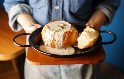 Fotografia de um chef segurando uma tábua de madeira, com uma panela grande preta em cima, com um pão aberto recheado com um creme de palmito que está transbordando e caindo na panela. Por cima, salsinha picada e salpicada.