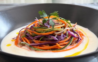 Fotografia mostra um prato fundo coberto com um molho de iogurte e macarrão de legumes coloridos.