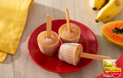 Picolés de banana e mamão em forminhas com palito, sobre prato vermelho, com frutas ao fundo, em mesa clara.