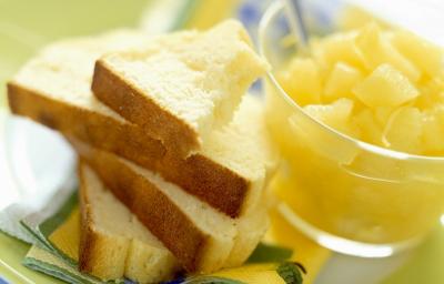 Fotografia em tons de amarelo, em uma bancada de madeira de cor branca. Ao centro, um prato verde contendo as fatias de pão com um recipiente ao lado contendo alguns pedaços de frutas. Fotografia em tons de amarelo, em uma bancada de madeira de cor branca. Ao centro, um prato verde contendo as fatias de pão com um recipiente ao lado contendo alguns pedaços de frutas.