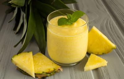 Fotografia de um copo de vidro com um gelado de abacaxi, e no topo, duas folhas de hortelã. Ao lado do copo tem alguns pedaços da fruta, sobre uma madeira em tons de cinza. Fotografia de um copo de vidro com um gelado de abacaxi, e no topo, duas folhas de hortelã. Ao lado do copo tem alguns pedaços da fruta, sobre uma madeira em tons de cinza.