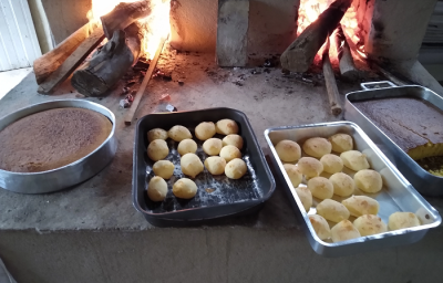 Foto em tons escuros da receita de pão de queijo servida em 30 porções divididas em duas formas sobre um fogão a lenha. Ao lado á mais duas formas com massas escuras e ao fundo estão dispostos diversos tocos de lenha