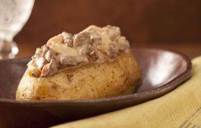 Fotografia em tons de marrom e pardo, em uma bancada contém um pano cor pardo com um garfo por cima, um recipiente marrom com uma batata com pedaços de carne com molho por cima e ao lado um copo transparente com agua Fotografia em tons de marrom e pardo, em uma bancada contém um pano cor pardo com um garfo por cima, um recipiente marrom com uma batata com pedaços de carne com molho por cima e ao lado um copo transparente com agua