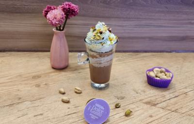 Fotografia em tons de lilás em uma bancada de madeira, com uma xícara de vidro e o Mochaccino Avelã com chantilly de pistache em cima para decorar. Em destaque, a cápsula de Dolce Gusto e ao fundo um vaso rosa com flores rosas. Fotografia em tons de lilás em uma bancada de madeira, com uma xícara de vidro e o Mochaccino Avelã com chantilly de pistache em cima para decorar. Em destaque, a cápsula de Dolce Gusto e ao fundo um vaso rosa com flores rosas.