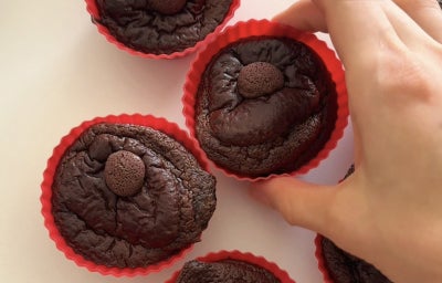 Fotografia mostra uma mão segurando um muffin de chocolate, fofinhos, com pedaço de chocolate derretido no topo, servidos em forminhas de silicone vermelha.