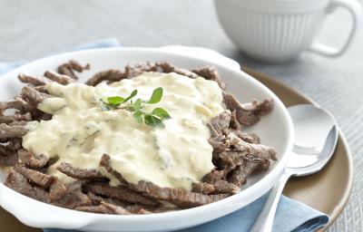 Fotografia em tons de branco e cinza, com carne em tiras e molho dentro de um prato branco sobre guardanapo azul e sous plat bege com colher ao lado, ao alto molheira branca, tudo sobre toalha cinza.
