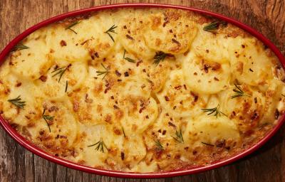 Foto bem aproximada de uma travessa vermelho dentro da qual está a receita de batatas gratinadas, decorada com ramos verdes de alecrim Foto bem aproximada de uma travessa vermelho dentro da qual está a receita de batatas gratinadas, decorada com ramos verdes de alecrim