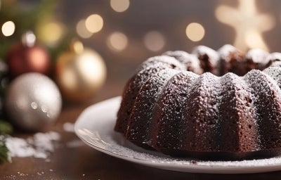 Fotografia de um bolo de chocolate em forma de bundt, em um prato branco, polvilhado com açúcar de confeiteiro. O fundo apresenta decorações natalinas em tons de ouro e prata.