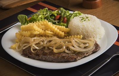 Fotografia em tons de marrom, branco e amarelo de uma bancada marrom, contém um prato redondo e branco com um bife com cebolas por cima, arroz, batatas fritas e salada como acompanhamento