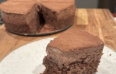 Fotografia de um bolo de chocolate caseiro, redondo e com uma cobertura de cacau. Uma fatia foi cortada, revelando seu interior úmido e fofo, apresentada em um prato branco, com um fundo de cozinha suave e acolhedor.