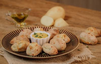 Fotografia em tons de marrom de uma bancada de madeira com um paninho de juta sobre ele um prato dourado redondo com croquetes de batata e ao centro um potinho redondo branco com molho. Ao fundo uma jarrinha com azeite e uma batata cortada. Fotografia em tons de marrom de uma bancada de madeira com um paninho de juta sobre ele um prato dourado redondo com croquetes de batata e ao centro um potinho redondo branco com molho. Ao fundo uma jarrinha com azeite e uma batata cortada.