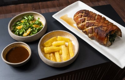 Pernil suíno assado com mandioca dourada, salada de folhas com laranja e molho servido à parte, apresentado em travessas.
