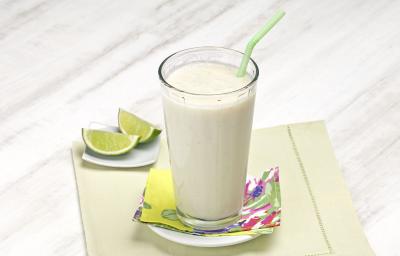 Fotografia em tons de verde em uma bancada de madeira cinza clara com um pano bege com detalhes verdes, um pratinho branco pequeno com um copo de vidro longo e grande com o smoothie de pera com limão. Ao lado, duas fatias de limão. Fotografia em tons de verde em uma bancada de madeira cinza clara com um pano bege com detalhes verdes, um pratinho branco pequeno com um copo de vidro longo e grande com o smoothie de pera com limão. Ao lado, duas fatias de limão.