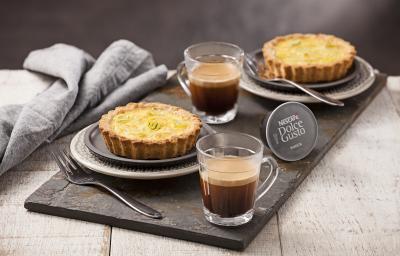 Fotografia em tons de cinza e dourado, ao centro dois copos com café, duas quiches assadas sobre  prato cinza e outro branco com borda decorada, com garfos ao lado, cápsula de café, sobre tábua grafite, guardanapo cinza, tudo sobre bancada em pátina.
