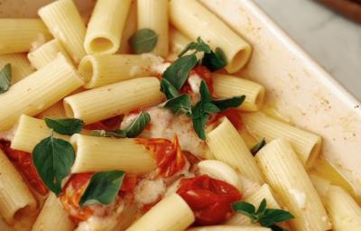 Fotografia em tons claros de uma travessa média, branca e retangular sobre uma mesa de mármore branca. Dentro da travessa tem uma receita de macarrão com tomate assado, manjericão, alhos inteiros assados e queijo. Fotografia em tons claros de uma travessa média, branca e retangular sobre uma mesa de mármore branca. Dentro da travessa tem uma receita de macarrão com tomate assado, manjericão, alhos inteiros assados e queijo.
