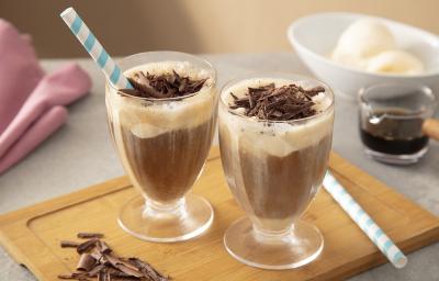 Foto em tons de marrom da receita de affogato de nescafé dolce gusto servida em duas xícaras de vidro dispostas sobre uma tábua de madeira com raspas de chocolate ao canto, além de canudos listrados em azul. Ao fundo uma xícara de café e sorvete.
