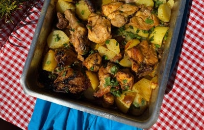 Frango assado dourado com batatas em rodelas levemente crocantes nas bordas, tudo coberto com cebolas caramelizadas e cheiro-verde fresco. Servido em assadeira rústica sobre mesa de madeira.