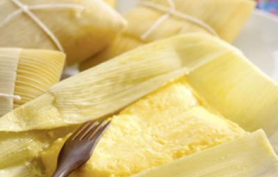 Fotografia em tons de amarelo em uma bancada de madeira, uma toalha colorida florida e algumas pamonhas de queijo coalho e alecrim em cima da bancada.
