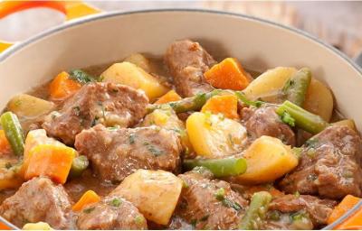 Fotografia em tons de laranja de uma bancada de madeira, com uma tábua de madeira, um pano quadriculado amarelo, vermelho e azul, uma colher prateada e uma panela laranja com alças com pedaços de carne, batata, cenoura e vagem.