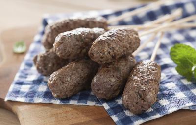 Fotografia em tons de azul em uma bancada de madeira com um pano xadrez azul e mini kaftas em palitos em cima dele. Ao lado, folhas de hortelã. Fotografia em tons de azul em uma bancada de madeira com um pano xadrez azul e mini kaftas em palitos em cima dele. Ao lado, folhas de hortelã.