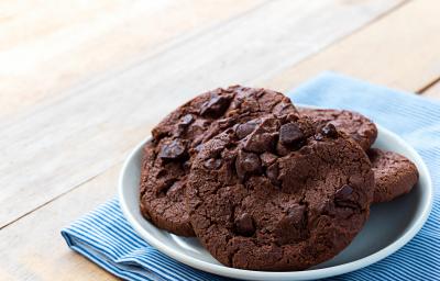Fotografia de quatro cookies de chocolate com gotas de chocolate dentro de um recipiente de vidro branco um pouco fundo. O recipiente está por cima de um paninho azul com listras brancas sobre uma mesa de madeira. Fotografia de quatro cookies de chocolate com gotas de chocolate dentro de um recipiente de vidro branco um pouco fundo. O recipiente está por cima de um paninho azul com listras brancas sobre uma mesa de madeira.