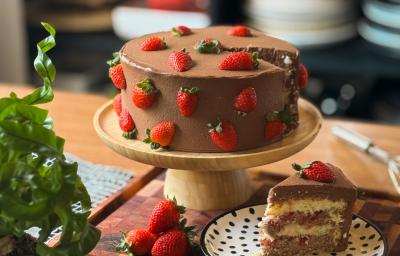 Fotografia em tons de marrom com um bolo de morango ao centro. O bolo possui massa branca e é recheado com camadas de recheio de chocolate e finalizado com morangos.