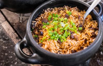 Fotografia em tons de marrom em uma bancada de madeira de cor marrom. Ao centro, uma panela preta contendo o arroz. Fotografia em tons de marrom em uma bancada de madeira de cor marrom. Ao centro, uma panela preta contendo o arroz.