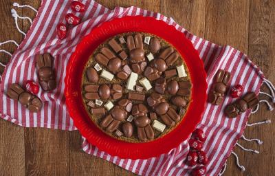 Fotografia em tons de vermelho, em uma bancada de madeira de cor marrom. Ao centro, uma toalha vermelha listrada com um prato vermelho em cima contendo o cookie em fatias. Ao redor do prato, há alguns KitKat's bunny e mini ovinhos de KitKat. Fotografia em tons de vermelho, em uma bancada de madeira de cor marrom. Ao centro, uma toalha vermelha listrada com um prato vermelho em cima contendo o cookie em fatias. Ao redor do prato, há alguns KitKat's bunny e mini ovinhos de KitKat.