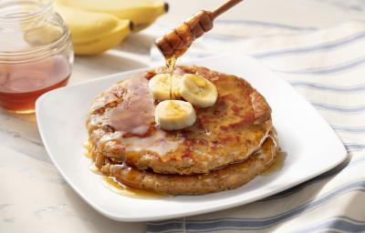 Fotografia me tons de branco de uma bancada de madeira, sobre ele um paninho listrado azul e branco com um prato quadrado com duas panquecas, mel e bananas. Ao fundo duas bananas e um recipiente de vidro com mel.