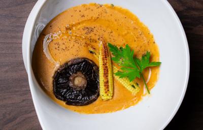 Fotografia em tons de branco com um prato branco ao centro. Em cima do prato existe uma receita de sopa de cor alaranjada com pedaços de cogumelo, mini milho e coentro em cima.