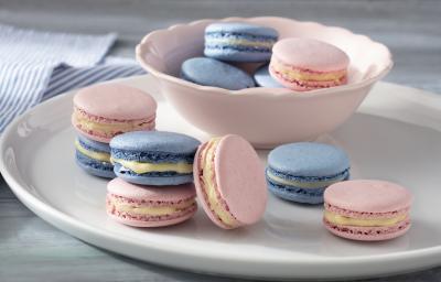 Fotografia em tons de azul e rosa em uma bancada de madeira branca clara com vários macarons coloridos em um prato branco e um paninho azul listrado.
