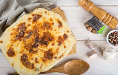Fotografia de uma travessa de vidro quadrada de bacalhau gratinado com batata sobre uma tábua de madeira e um pano de tom cinza. No fundo tem um moedor de pimenta de madeira.