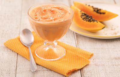 Foto em tons de laranja da receita de creme de papaia servida em uma taça de vidro sobre um pano laranja em uma mesa de madeira. Ao lado há um colher prateada e ao fundo há um prato com duas fatias médias de mamão Foto em tons de laranja da receita de creme de papaia servida em uma taça de vidro sobre um pano laranja em uma mesa de madeira. Ao lado há um colher prateada e ao fundo há um prato com duas fatias médias de mamão