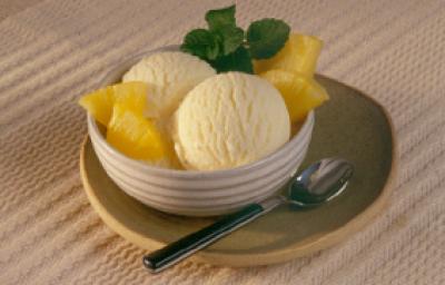 Fotografia com tons de bege e amarelo, ao centro bowl com duas bolas de sorvete de abacaxi, abacaxi fatiado, hortelão, sobre prato verde, ao lado uma colher de sobremesa, sobre toalha bege. Fotografia com tons de bege e amarelo, ao centro bowl com duas bolas de sorvete de abacaxi, abacaxi fatiado, hortelão, sobre prato verde, ao lado uma colher de sobremesa, sobre toalha bege.