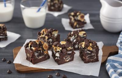 Foto da receita de Brownie de Negresco Coberto, em pedaços, sobre uma tábua de madeira em um balcão decorado com alguns copos com leite, um tecido azul listrado e algumas gotas de chocolate