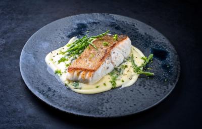 Fotografia um filé de pescada assado sobre um creme de legumes em um prato raso escuro, o qual está sobre uma mesa preta.